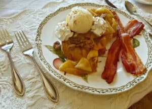 French toast topped with peaches and whipped cream with bacon on china plate.