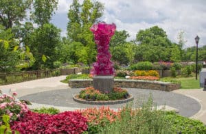 Pink glass sculpture on a plinth in a garden filled with pink and green plants.