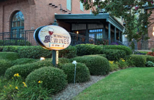 Large brick building with green trim surrounded by greenery and a wooden sign with a wine glass and grapes.