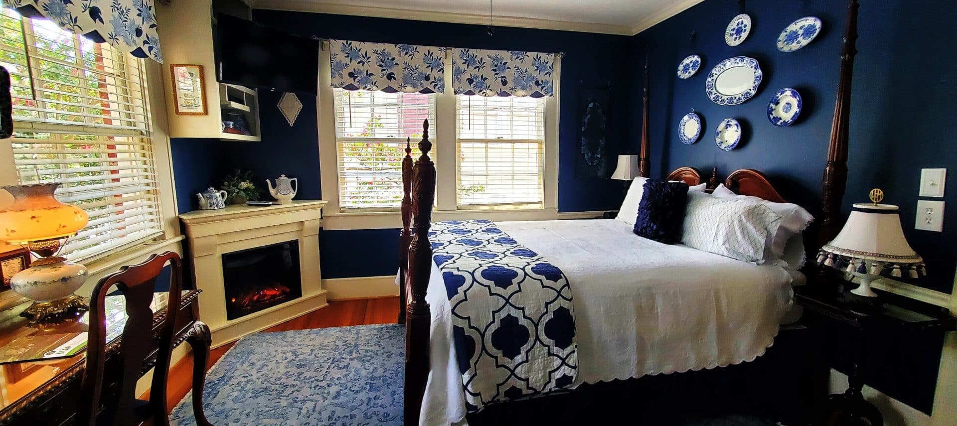 Four poster queen bed in white and blue with windows and fire place.
