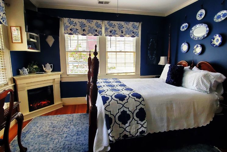 Four poster queen bed in white and blue with windows and fire place.