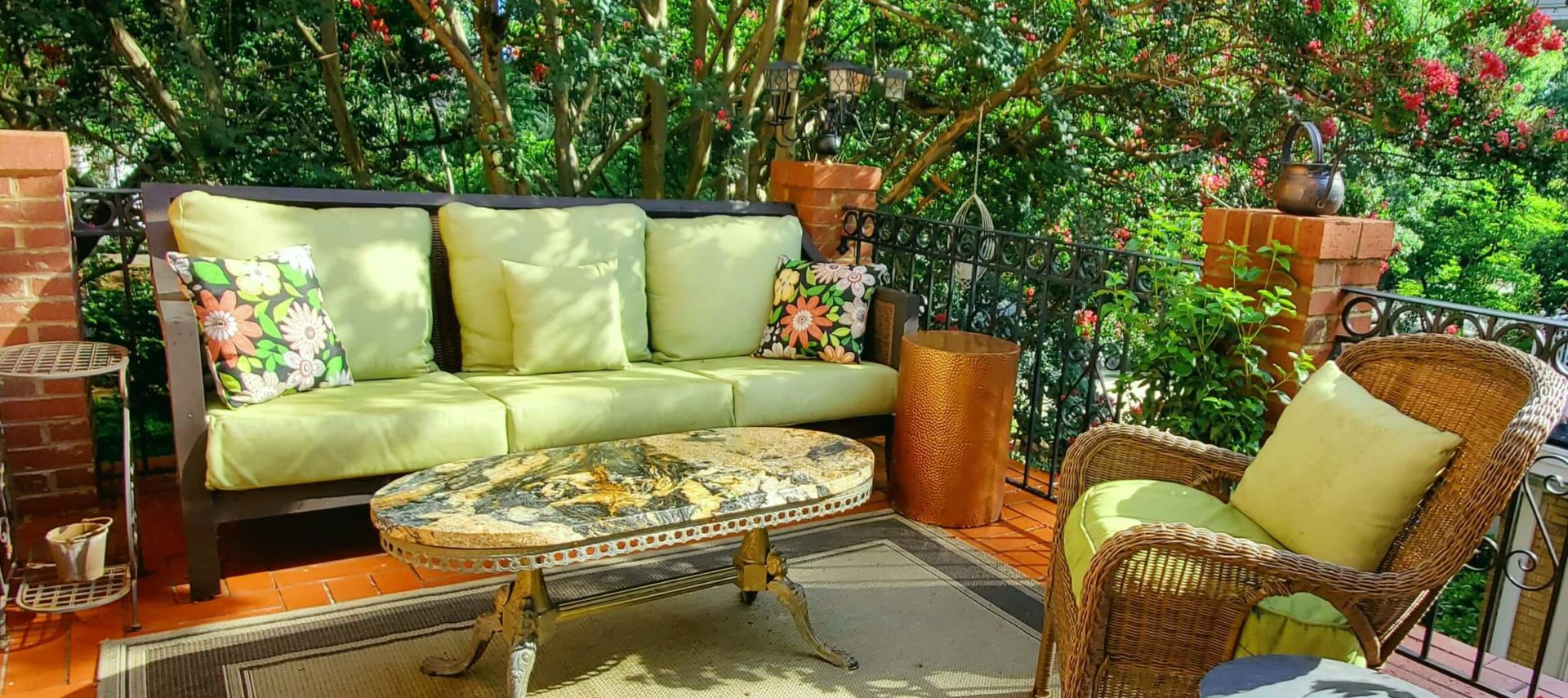 Outdoor couch and wicker chair on bricked balcony with marble table with Crepe myrtle tree in bloom.