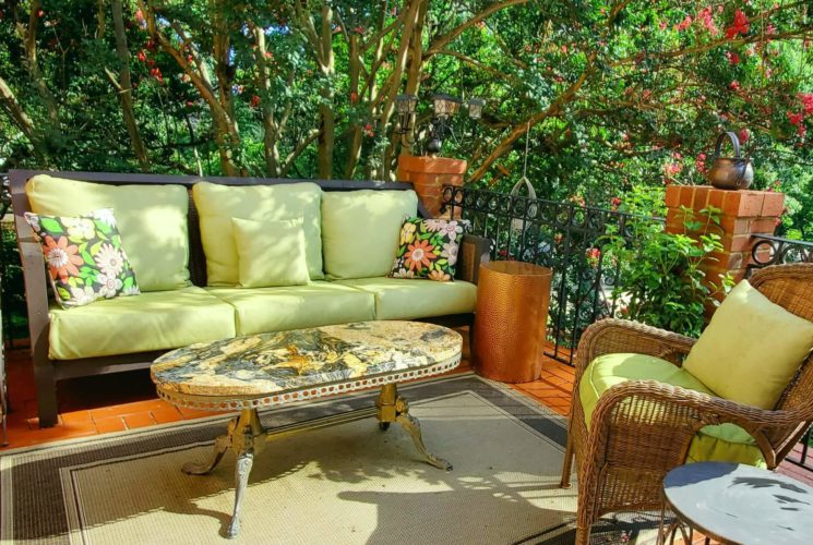 Outdoor couch and wicker chair on bricked balcony with marble table with Crepe myrtle tree in bloom.