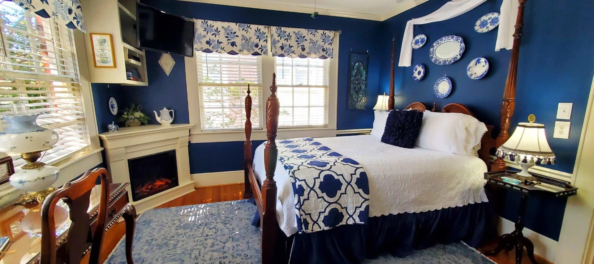 Queen bed with white bed spread in blue room with lit fireplace.