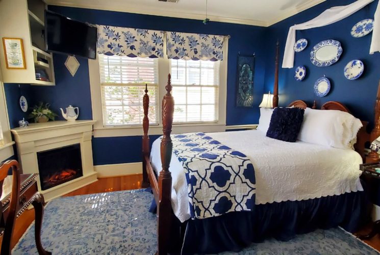 Queen bed with white bed spread in blue room with lit fireplace.