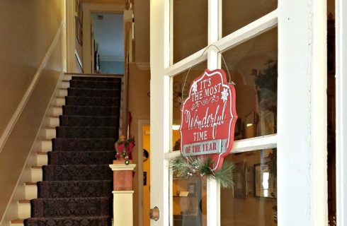 Open French door with red Christmas sign showing a tall carpeted staircase leading to the upstairs of a home