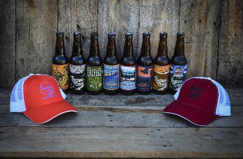 Two ballcaps on a wooden bench in front of a line of 8 different beer bottles.