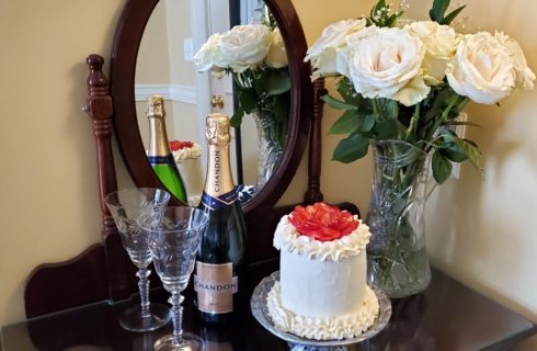 Strawberry and buttercream champagne cake with white roses, champagne, and cut glass flutes on mirrored desk.