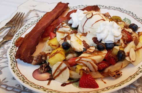 French toast covered with bananas, strawberries, pineapple, blueberries, and whipped cream. accompanied by bacon.