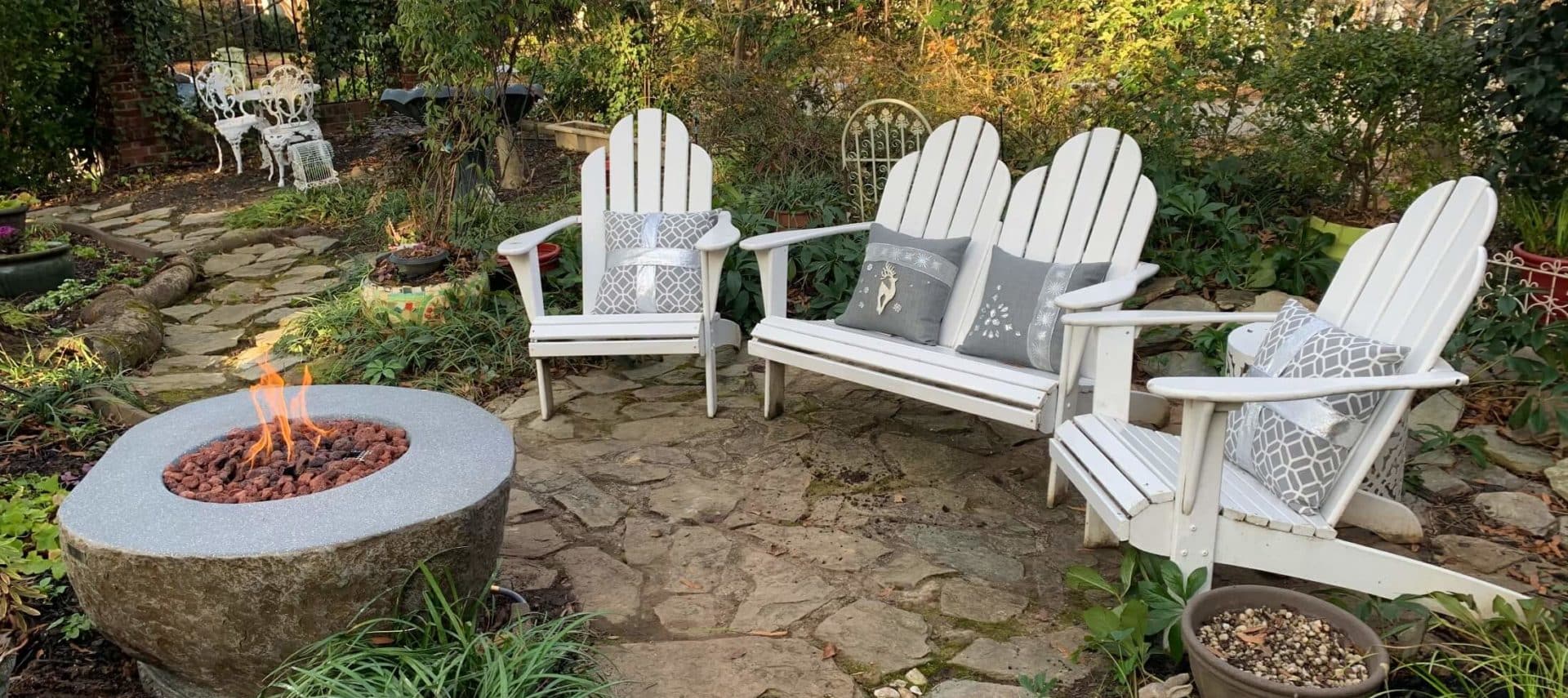 White Adirondack chairs with gray cushions surrounding stone fire table in English garden.