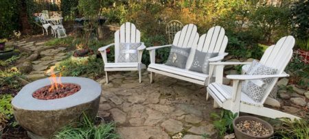 White Adirondack chairs with gray cushions surrounding stone fire table in English garden.
