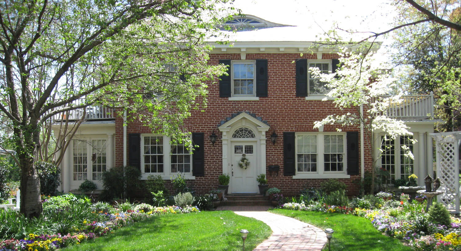 Large brick Colonial-style house fronted by flowering trees and green lawn. 