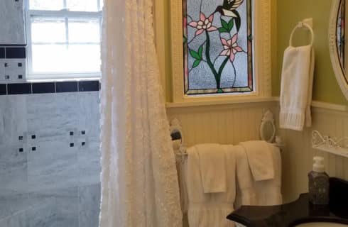 Bathroom in white with lace shower curtain and stained glass window.