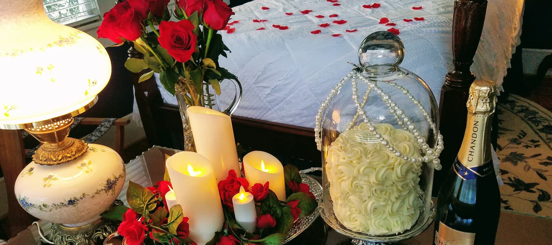 Side table in bedroom with candles, a small cake in a pearl-draped glass dome and chocolate covered strawberries.