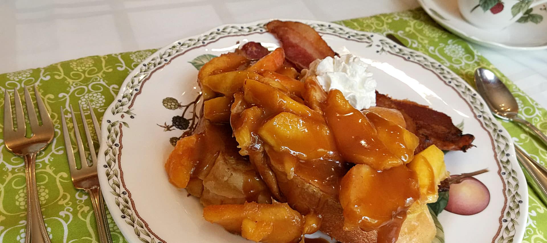 French toast topped with peaches and whipped cream with bacon on china plate.