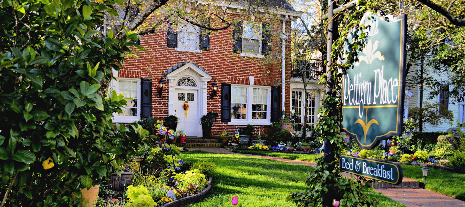 Large brick house with black shutters fronted by garden blooming with trees and flowers.