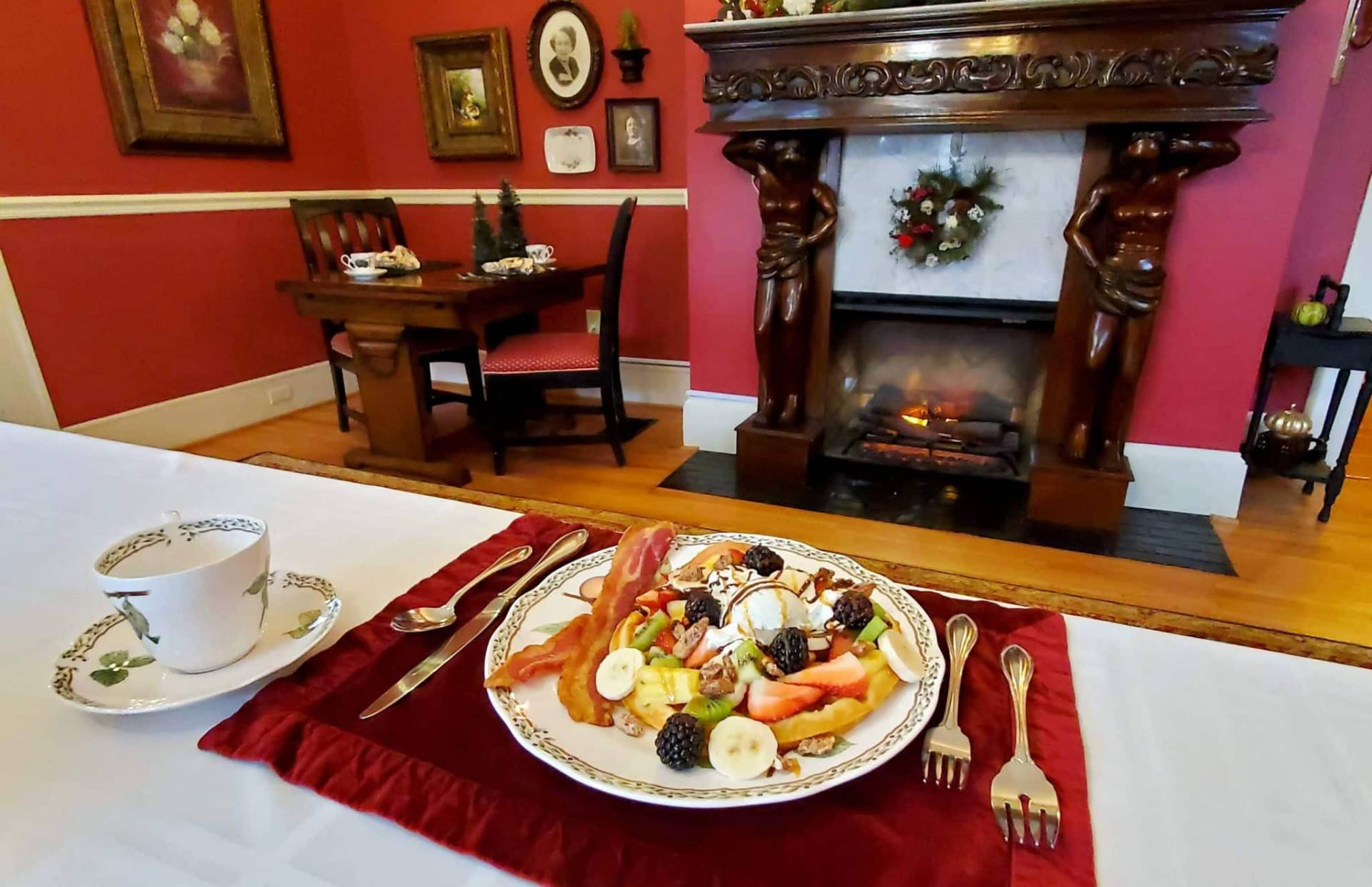 Banana split waffle covered in fruit with bacon on set table in front of fireplace. 