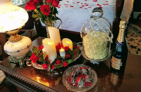 Side table in bedroom with romantic package including candles, a small cake in a pearl-draped glass dome and chocolate covered strawberries.